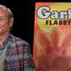Bill Murray wears a plaid suit jacket, sweater vest and striped shirt while sitting next to a poster for the fake Garfield Flabby Tabby 3 film as seen in Zombieland: Double Tap.